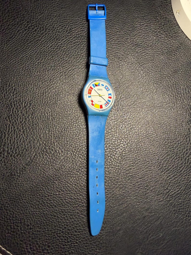 K&ouml;nnte beinhalten: Eine blaue analoge Armbanduhr mit einem runden wei&szlig;en Zifferblatt, das bunte nautische Signalflaggen als Stundenmarkierungen zeigt. Die Uhr hat ein passendes blaues Silikonarmband mit Dornschlie&szlig;e sowie goldfarbene Zeiger.