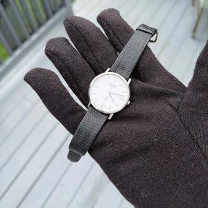 May include: A silver-tone analogue wristwatch featuring a round white dial with thin black hour markers and hands. The watch includes a textured black leather strap with a silver buckle, displaying the Seiko Quartz logo on the watch face.