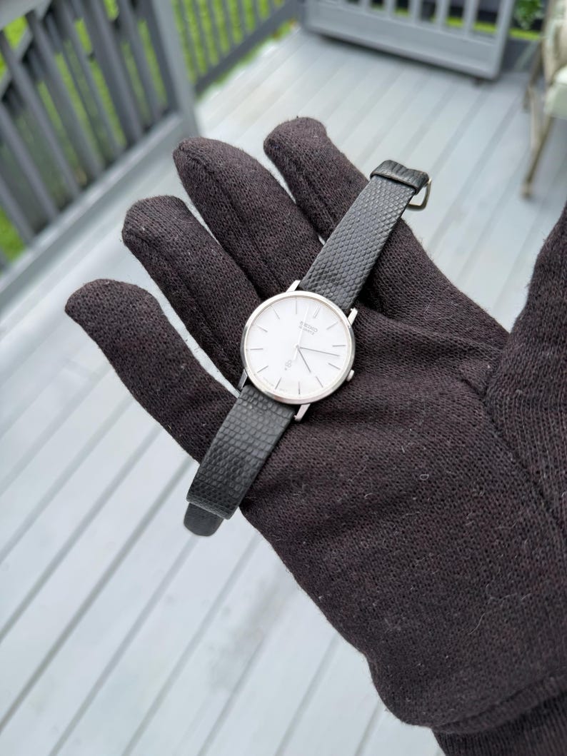 May include: A silver-tone analogue wristwatch featuring a round white dial with thin black hour markers and hands. The watch includes a textured black leather strap with a silver buckle, displaying the Seiko Quartz logo on the watch face.