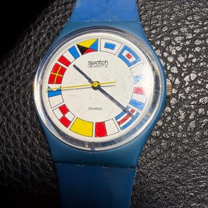K&ouml;nnte beinhalten: Eine blaue Swatch Quartz Armbanduhr mit wei&szlig;em Zifferblatt, das einen kreisf&ouml;rmigen Rand aus bunten nautischen Signalflaggen aufweist. Die Uhr hat ein blaues Silikonarmband, einen gelben Sekundenzeiger und schwarze Zeiger. Das Wort SWISS ist aufgedruckt.