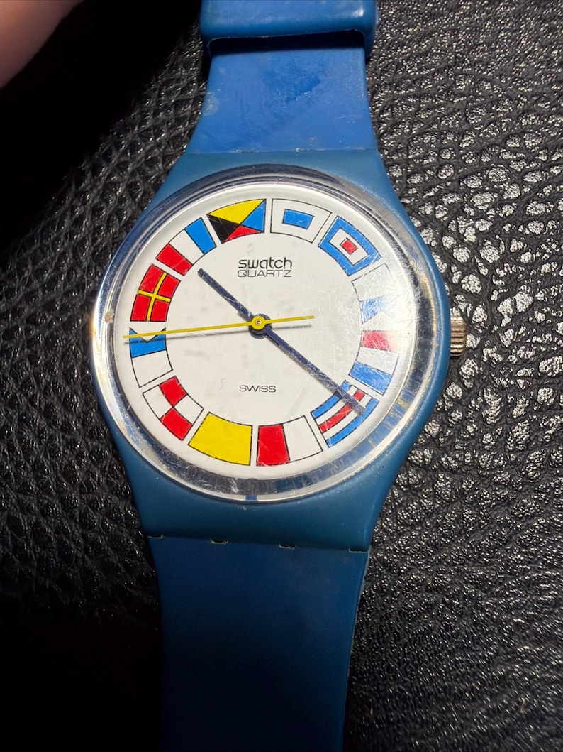 K&ouml;nnte beinhalten: Eine blaue Swatch Quartz Armbanduhr mit wei&szlig;em Zifferblatt, das einen kreisf&ouml;rmigen Rand aus bunten nautischen Signalflaggen aufweist. Die Uhr hat ein blaues Silikonarmband, einen gelben Sekundenzeiger und schwarze Zeiger. Das Wort SWISS ist aufgedruckt.