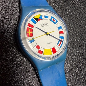K&ouml;nnte beinhalten: Eine blaue Swatch Quartz Armbanduhr mit einem wei&szlig;en Zifferblatt, das am Rand mit bunten nautischen Signalflaggen verziert ist. Die Uhr hat ein blaues Kunststoffarmband, ein klares rundes Geh&auml;use sowie schmale Stunden-, Minuten- und Sekundenzeiger.