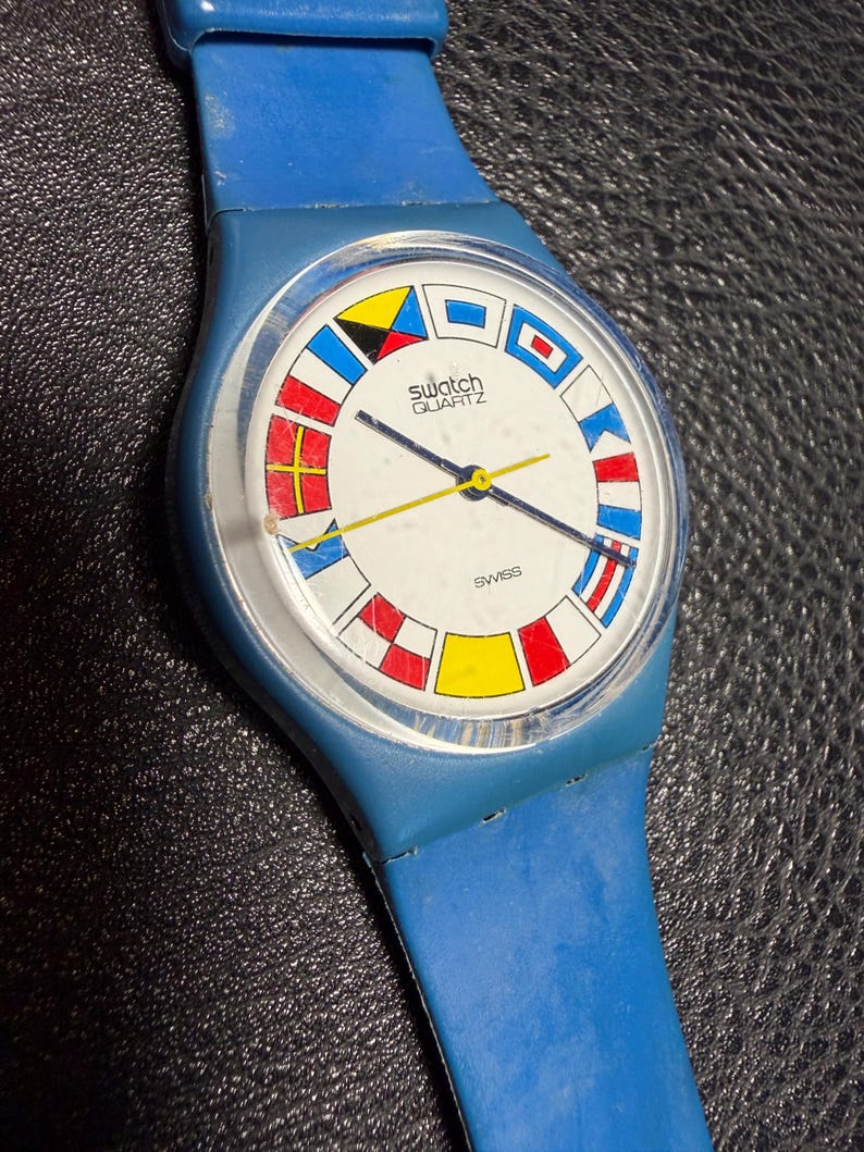 K&ouml;nnte beinhalten: Eine blaue Swatch Quartz Armbanduhr mit einem wei&szlig;en Zifferblatt, das am Rand mit bunten nautischen Signalflaggen verziert ist. Die Uhr hat ein blaues Kunststoffarmband, ein klares rundes Geh&auml;use sowie schmale Stunden-, Minuten- und Sekundenzeiger.