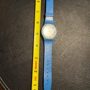 K&ouml;nnte beinhalten: Eine blaue Armbanduhr mit einem runden Zifferblatt, das nautische Signalflaggen als Stundenmarkierungen aufweist. Die Uhr liegt neben einem gelben Ma&szlig;band, das eine Gesamtl&auml;nge von etwa 23 cm von der Schnalle bis zum Bandende anzeigt.
