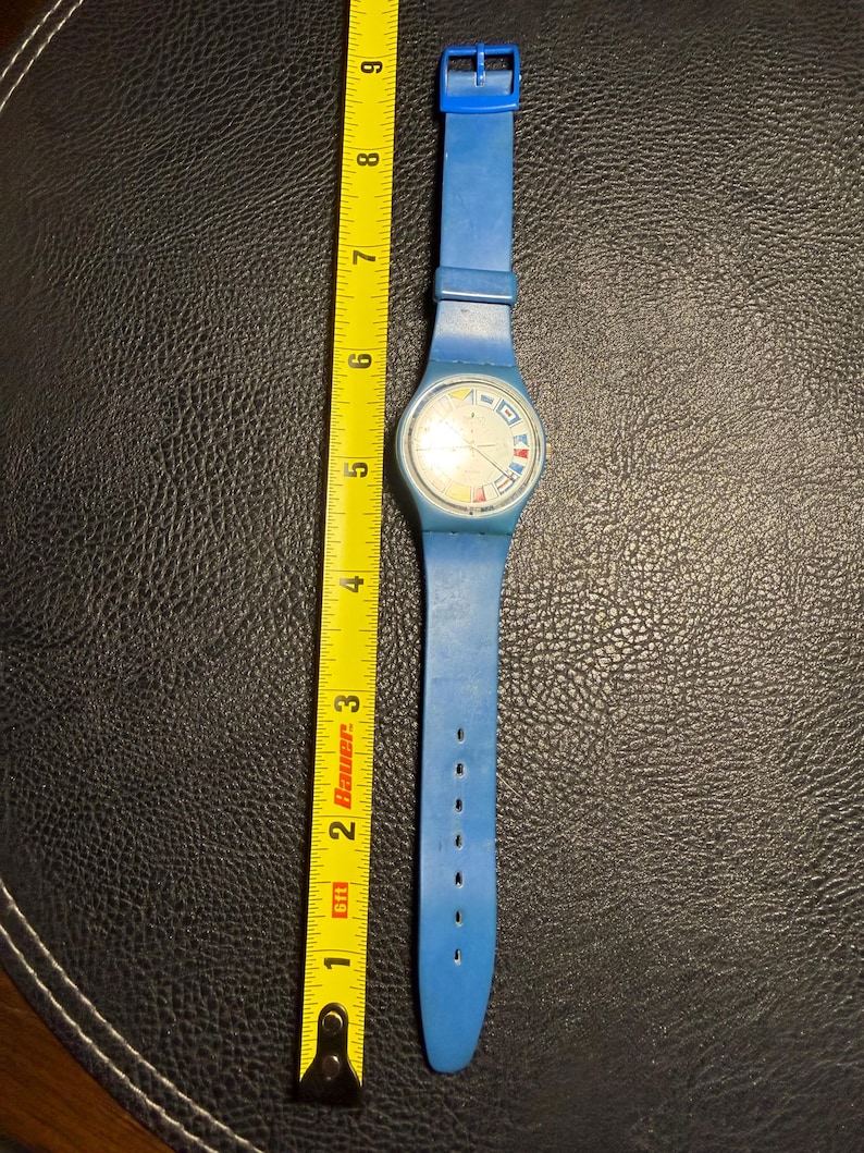 K&ouml;nnte beinhalten: Eine blaue Armbanduhr mit einem runden Zifferblatt, das nautische Signalflaggen als Stundenmarkierungen aufweist. Die Uhr liegt neben einem gelben Ma&szlig;band, das eine Gesamtl&auml;nge von etwa 23 cm von der Schnalle bis zum Bandende anzeigt.