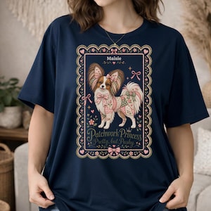 May include: A navy blue t-shirt featuring a rectangular graphic with a scalloped border. The design shows a Papillon dog with patchwork patterns, bows, and flowers. Text reads Maisie, Patchwork Princess, and Pretty, but Pushy.