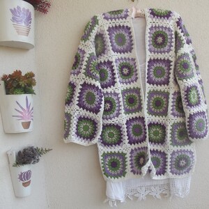 May include: A long-sleeved cardigan made of joined crochet granny squares in shades of purple, green, and off-white. The garment features a repeating circular floral pattern within each square, an open front design, and a white lace trim at the hem.