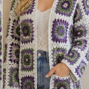 May include: A long-sleeved cardigan made of crocheted granny squares in shades of purple, green, and white. The garment features an open front design, a straight hem, and two front patch pockets, constructed from a repeating floral-inspired pattern.