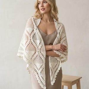 Puede incluir: Un chal de ganchillo color crema con un intrincado patrón geométrico de motivos triangulares y un borde festoneado. El chal está drapeado sobre un vestido lencero beige liso, mostrando la textura calada y el detallado trabajo del hilo.
