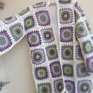 May include: A long-sleeved cardigan constructed from joined crochet granny squares. Each square features a circular floral pattern in shades of purple, lavender, and green, framed by a white border. The garment has an open front design.
