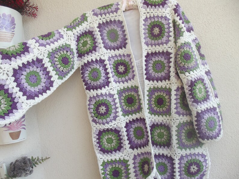 May include: A long-sleeved cardigan constructed from joined crochet granny squares. Each square features a circular floral pattern in shades of purple, lavender, and green, framed by a white border. The garment has an open front design.