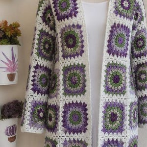 May include: A long-sleeved cardigan constructed from joined crochet granny squares in shades of purple, green, and white. The garment features an open front design with a white border and is displayed on a mannequin wearing a white undergarment.