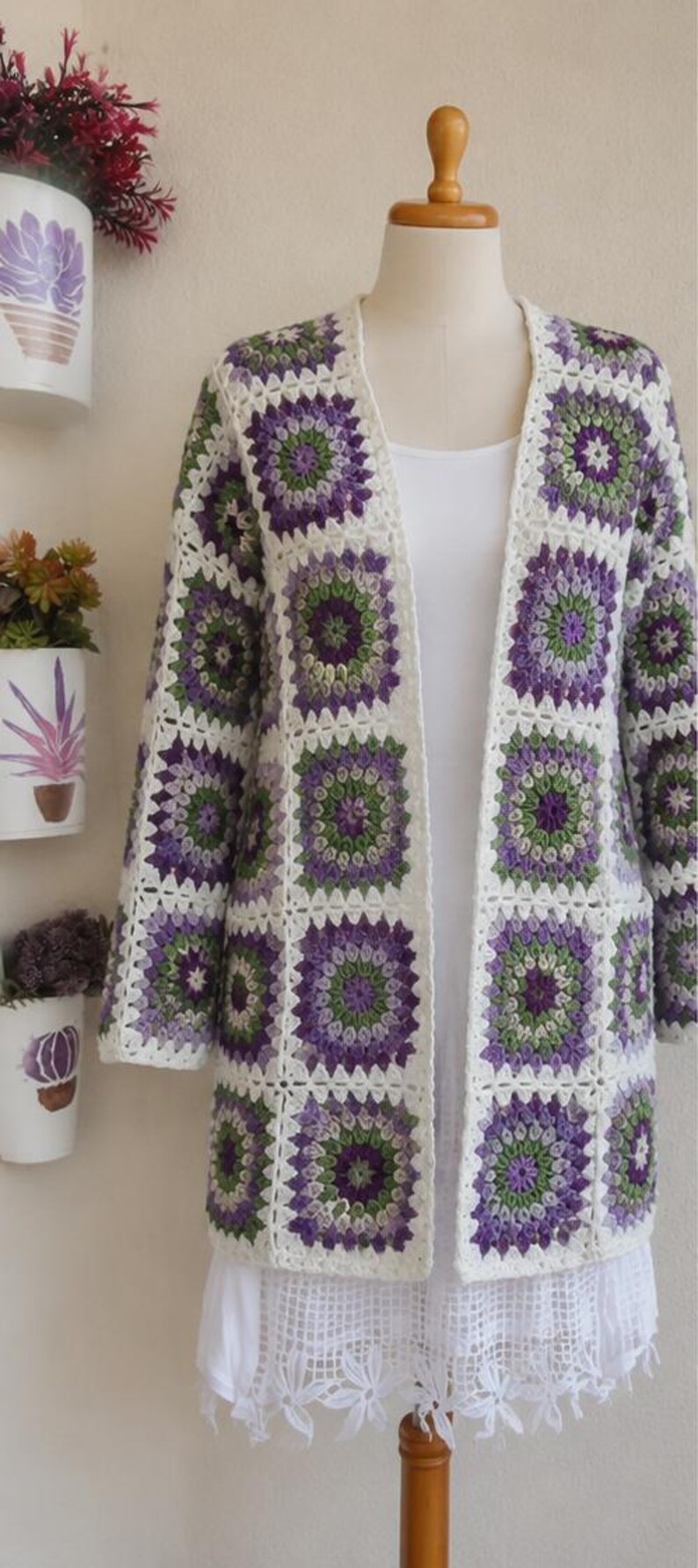 May include: A long-sleeved cardigan constructed from joined crochet granny squares in shades of purple, green, and white. The garment features an open front design with a white border and is displayed on a mannequin wearing a white undergarment.