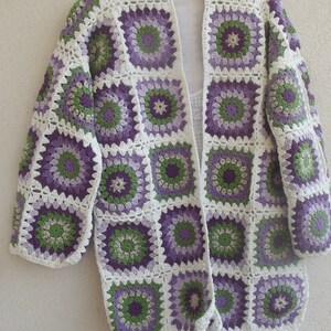 May include: A long-sleeved cardigan constructed from joined crochet granny squares. The squares feature a circular floral pattern in shades of purple, light purple, and green, all bordered by a white yarn frame. The garment has an open front.
