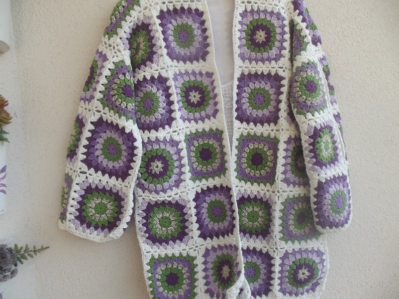 May include: A long-sleeved cardigan constructed from joined crochet granny squares. The squares feature a circular floral pattern in shades of purple, light purple, and green, all bordered by a white yarn frame. The garment has an open front.