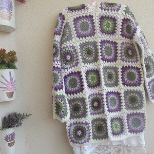 May include: A long-sleeved cardigan made of crocheted granny squares in shades of purple, light purple, green, and white. The garment features a repeating circular floral pattern within each square, joined by white yarn, with a white hem.