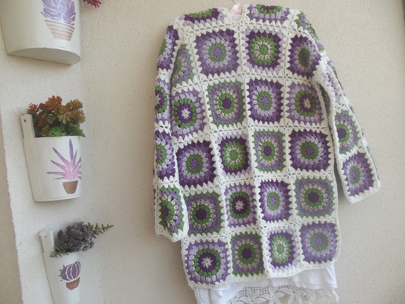 May include: A long-sleeved cardigan made of crocheted granny squares in shades of purple, light purple, green, and white. The garment features a repeating circular floral pattern within each square, joined by white yarn, with a white hem.