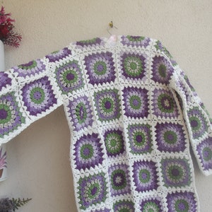 May include: A long-sleeved cardigan constructed from joined crochet granny squares. The squares feature a circular floral pattern in shades of purple, light green, and dark green, all bordered by a white yarn frame that connects the pieces.