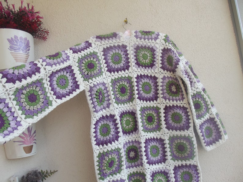 May include: A long-sleeved cardigan constructed from joined crochet granny squares. The squares feature a circular floral pattern in shades of purple, light green, and dark green, all bordered by a white yarn frame that connects the pieces.