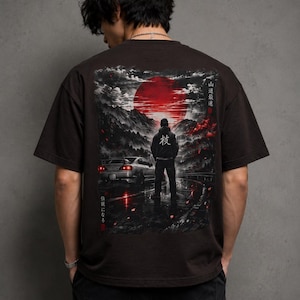 May include: A dark brown t-shirt featuring a graphic of a person standing in front of a car with a red sun and mountain landscape. Japanese text is visible on the shirt. The person is wearing a black hoodie. The shirt has short sleeves.