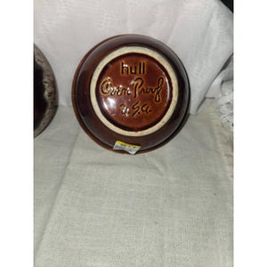 May include: A round brown ceramic bowl with a glossy finish. The underside features embossed text reading hull, Oven Proof, and U.S.A. A small white and yellow sticker is attached to the bottom edge of the bowl, which is positioned on a textured fabric.