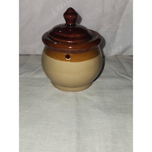 May include: A small, round ceramic jar with a glossy brown lid and a matching brown rim. The main body of the jar is a light beige color. It features a pointed, knob-like handle on top of the lid and a smooth, rounded base for stability.