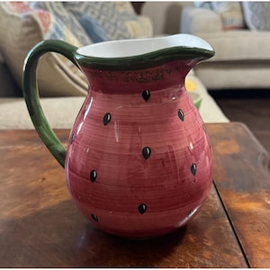 Peut inclure: Un pichet en céramique au corps arrondi peint dans des tons de rose et de rouge avec un effet de coups de pinceau texturés. L'extérieur est orné de petites graines noires en forme de goutte. Il possède une anse vert foncé et un intérieur blanc.