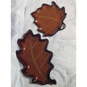 May include: Two glossy, dark brown ceramic dishes shaped like leaves. One dish is shaped like a maple leaf, while the other is shaped like an elongated oak leaf. Both dishes feature embossed vein details and a smooth, reflective glazed finish.