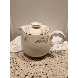 May include: A cream-colored ceramic teapot featuring a rounded body, a matching lid with a small knob, and a curved handle. The teapot is decorated with delicate gold-toned wheat stalk illustrations on both the lid and the main body surface.