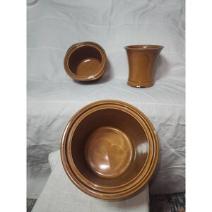 May include: Three pieces of brown glazed ceramic kitchenware including two round bowls with wide rims and one flared vase. The items feature a smooth, glossy finish and a consistent warm brown color throughout their surfaces.
