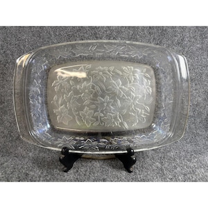 May include: A clear rectangular glass serving dish featuring an embossed floral pattern of poinsettia-like flowers and leaves across the center and along the wide, integrated handles on both sides, resting on a black plastic display stand.
