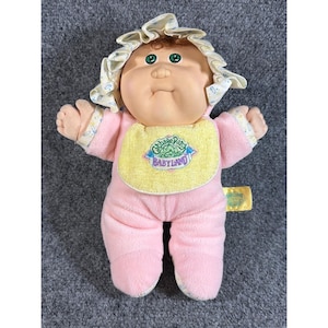 Könnte beinhalten: Eine Plüschpuppe mit rundem Gesicht, grünen Augen und braunen Haaren, die eine gerüschte Haube mit blau-gelbem Muster trägt. Die Puppe ist in einen rosa Fleece-Strampler mit einem gelben Lätzchen gekleidet, das den Text Cabbage Patch Babyland zeigt.