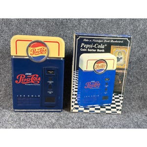 May include: A blue and yellow Pepsi-Cola coin sorter bank with a vintage vending machine design, featuring the Pepsi-Cola logo, the text Drink Pepsi-Cola, and Ice Cold. It is displayed next to its original cardboard packaging box.