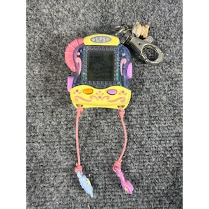 Könnte beinhalten: Ein violett-gelbes digitales Haustier-Spielzeug mit einem LPS-Logo auf dem oberen Rahmen. Das Gerät verfügt über einen Bildschirm mit einer kleinen Figur, zwei Tasten und einen Metall-Schlüsselanhänger. Zwei rosa Schnüre hängen unten.