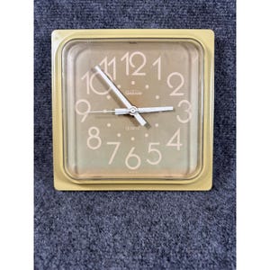 May include: A square wall clock with a light yellow plastic frame and a clear protective cover. The clock face features a light tan background with large white numerals from one to twelve, white clock hands, and the text Sunbeam and QUARTZ.