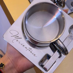 May include: A silver-colored stainless steel cup and saucer set with a polished finish. The square saucer features the name Mariantonia engraved in a cursive script. A matching metal spoon is included, resting in a dedicated slot on the saucer.
