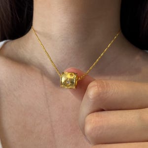 Könnte beinhalten: Eine goldfarbene Halskette mit einem zylindrischen Anhänger in matter, texturierter Optik und einem kleinen, klaren, rund geschliffenen Stein in der Mitte. Der Anhänger hängt an einer feinen, goldfarbenen Gliederkette.