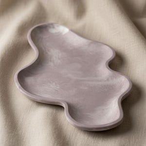 May include: A lavender-colored tray with an irregular, organic, cloud-like shape. The surface features a subtle, marbled texture with swirling patterns of lighter and darker shades of purple throughout the smooth, shallow basin of the tray.