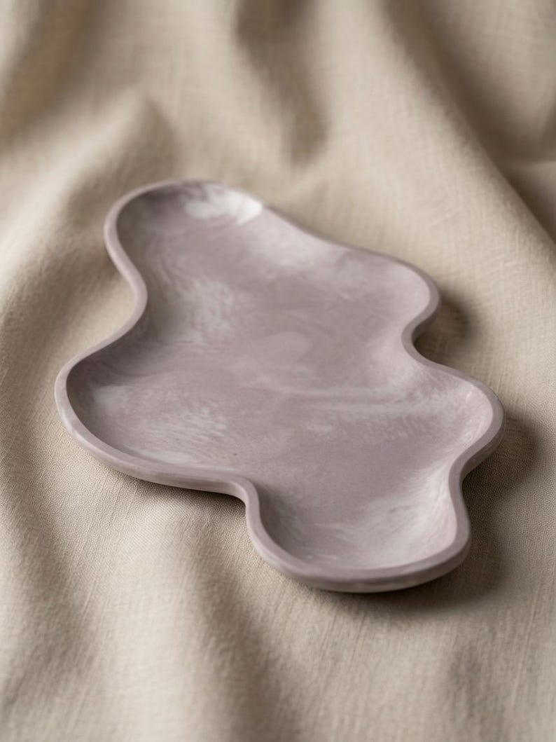 May include: A lavender-colored tray with an irregular, organic, cloud-like shape. The surface features a subtle, marbled texture with swirling patterns of lighter and darker shades of purple throughout the smooth, shallow basin of the tray.