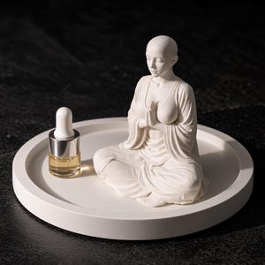 May include: A white circular tray holding a small glass dropper bottle with a gold collar and a white cap, alongside a white figurine of a person with a shaved head sitting in a meditative pose with hands pressed together in front of the chest.