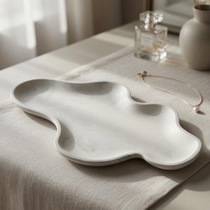 May include: An elongated, cloud-shaped tray featuring a smooth, white surface with subtle grey marble-like veining. The tray has a low, raised rim and a fluid, organic silhouette, providing a flat, open area for holding small items.