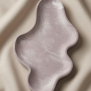May include: An irregular, organic-shaped tray featuring a smooth, matte finish in a soft, muted mauve color. The surface displays a subtle, swirling marbled pattern in lighter shades of white and pale pink, creating a fluid, cloud-like appearance.
