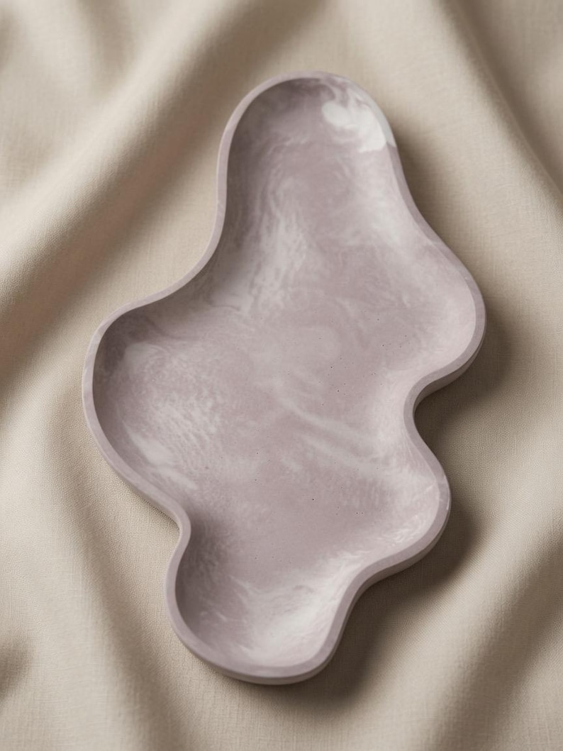 May include: An irregular, organic-shaped tray featuring a smooth, matte finish in a soft, muted mauve color. The surface displays a subtle, swirling marbled pattern in lighter shades of white and pale pink, creating a fluid, cloud-like appearance.