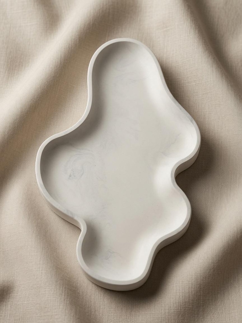 May include: An off-white tray with an irregular, organic, wavy shape and a subtle marbled texture. The surface is smooth and features soft, light gray swirls throughout the material, creating a distinct pattern within the curved edges.