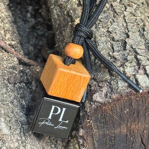 May include: A square black glass bottle with a light brown wooden cap and a black cord featuring a small wooden bead. The front of the bottle displays the white text PL and Pauline Lumière. The object is designed for hanging with a black cord.