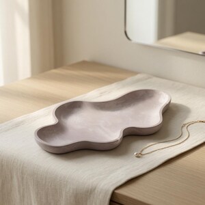 May include: A pale lavender, cloud-shaped decorative tray with a smooth, matte finish and undulating, organic edges. The tray features a shallow, recessed basin and a solid, continuous form, presented as a singular piece for holding small items.