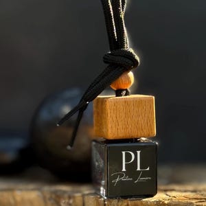 May include: A small square glass car air freshener bottle with a light brown wooden cap and a black cord for hanging. The front of the bottle features a black label with the white text PL and the script text Pauline Lumière printed below it.