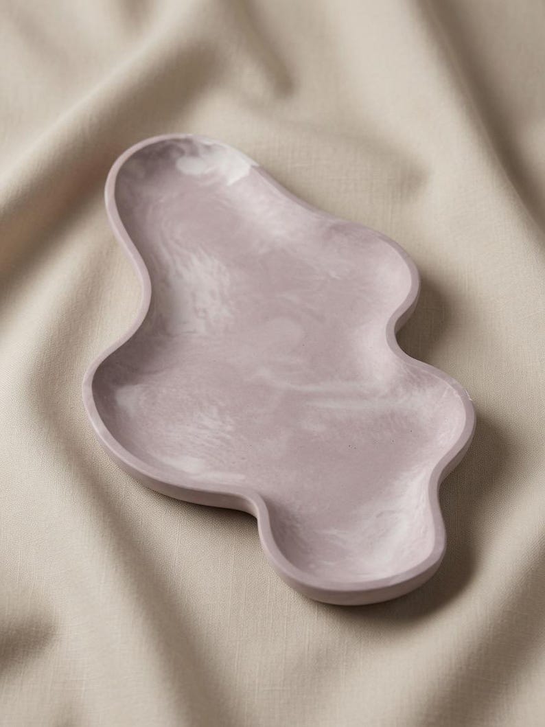 May include: An irregular, organic-shaped tray featuring a smooth, matte finish in a soft, muted lavender color with subtle, swirling white marbled patterns throughout the surface, creating a unique, flowing silhouette with raised, rounded edges.