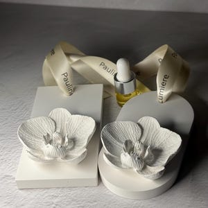 Könnte beinhalten: Zwei weiße Keramik-Orchideen-Ornamente, eines rechteckig und eines bogenförmig, jeweils mit detailliertem Blumenrelief. Eine kleine Glas-Tropfflasche mit silbernem Verschluss steht dazwischen, begleitet von einem cremefarbenen Band mit dem Aufdruck Pauline Lumiere.