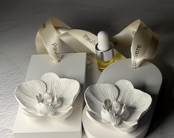 May include: Two white ceramic orchid ornaments, one rectangular and one arched, each featuring a detailed floral relief. A small glass dropper bottle with a silver cap sits between them, accompanied by a cream-colored ribbon printed with the text Pauline Lumiere.